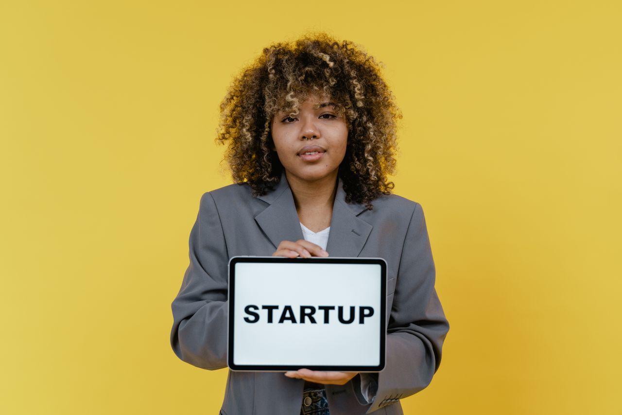 why-choose-us Portrait of a woman holding a tablet showing the word 'STARTUP' against a yellow background.